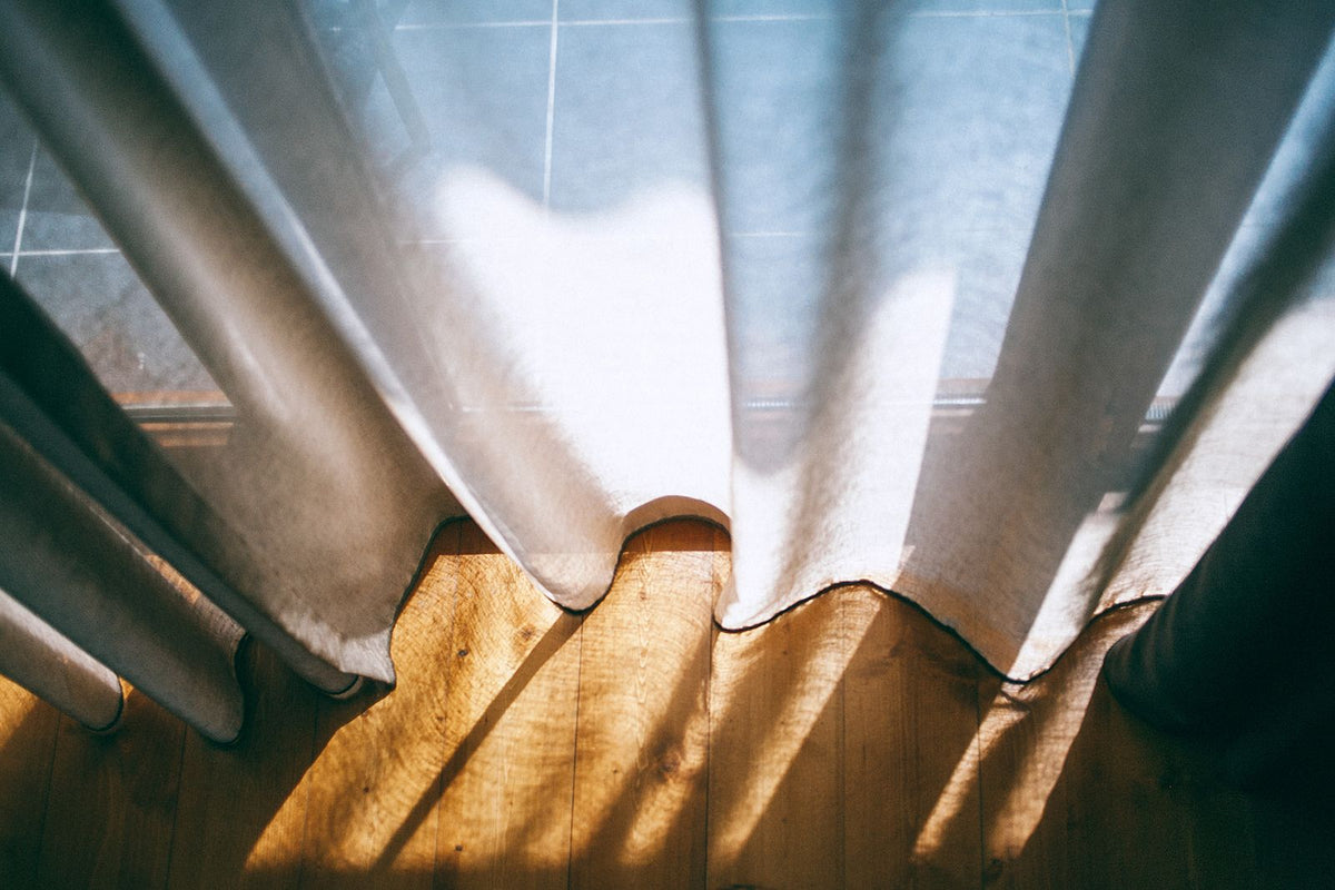 Rideaux clairs devant une fenêtre et raies de soleil sur le parquet d'un salon que l'on aère contre une odeur de tabac froid dans un appartement